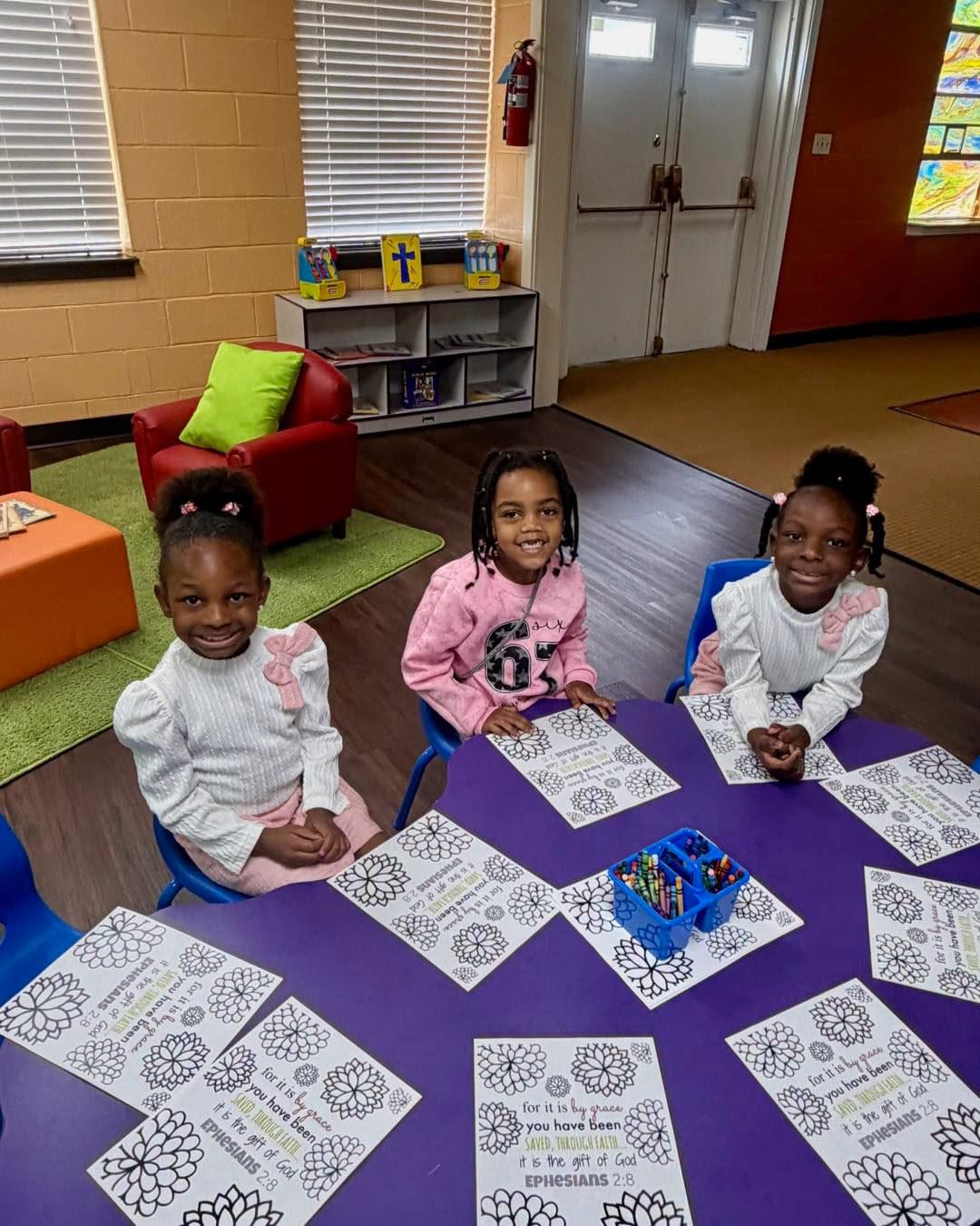 Three girls smiling at a purple table with Ephesians 2:8 coloring sheets and crayons in children's church.
