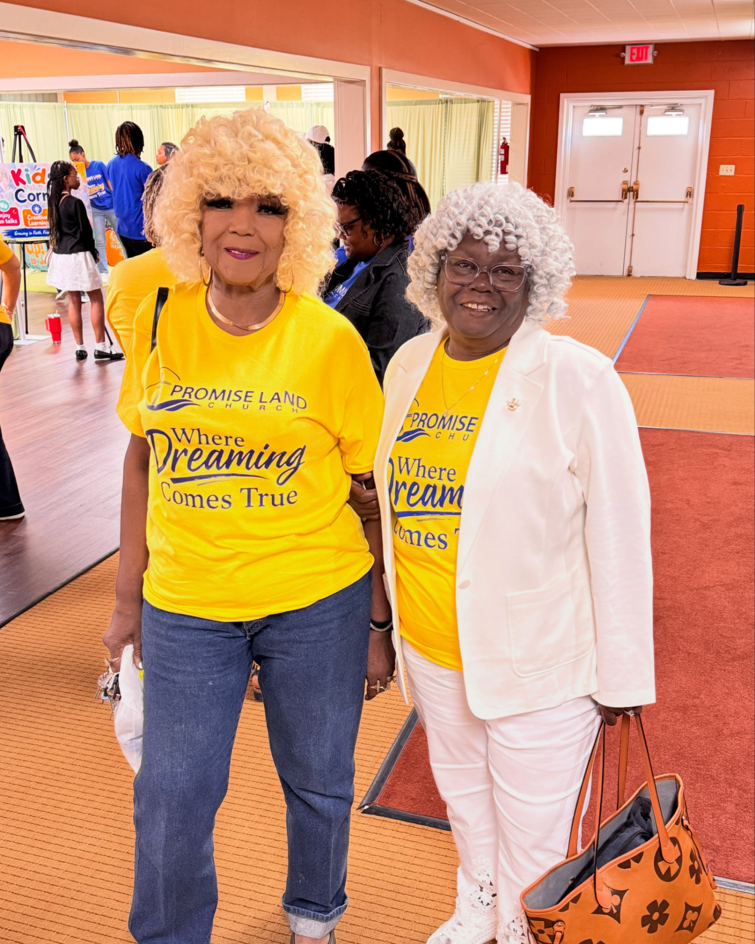 Two church mothers in yellow Promise Land Church shirts standing arm in arm at a church gathering.