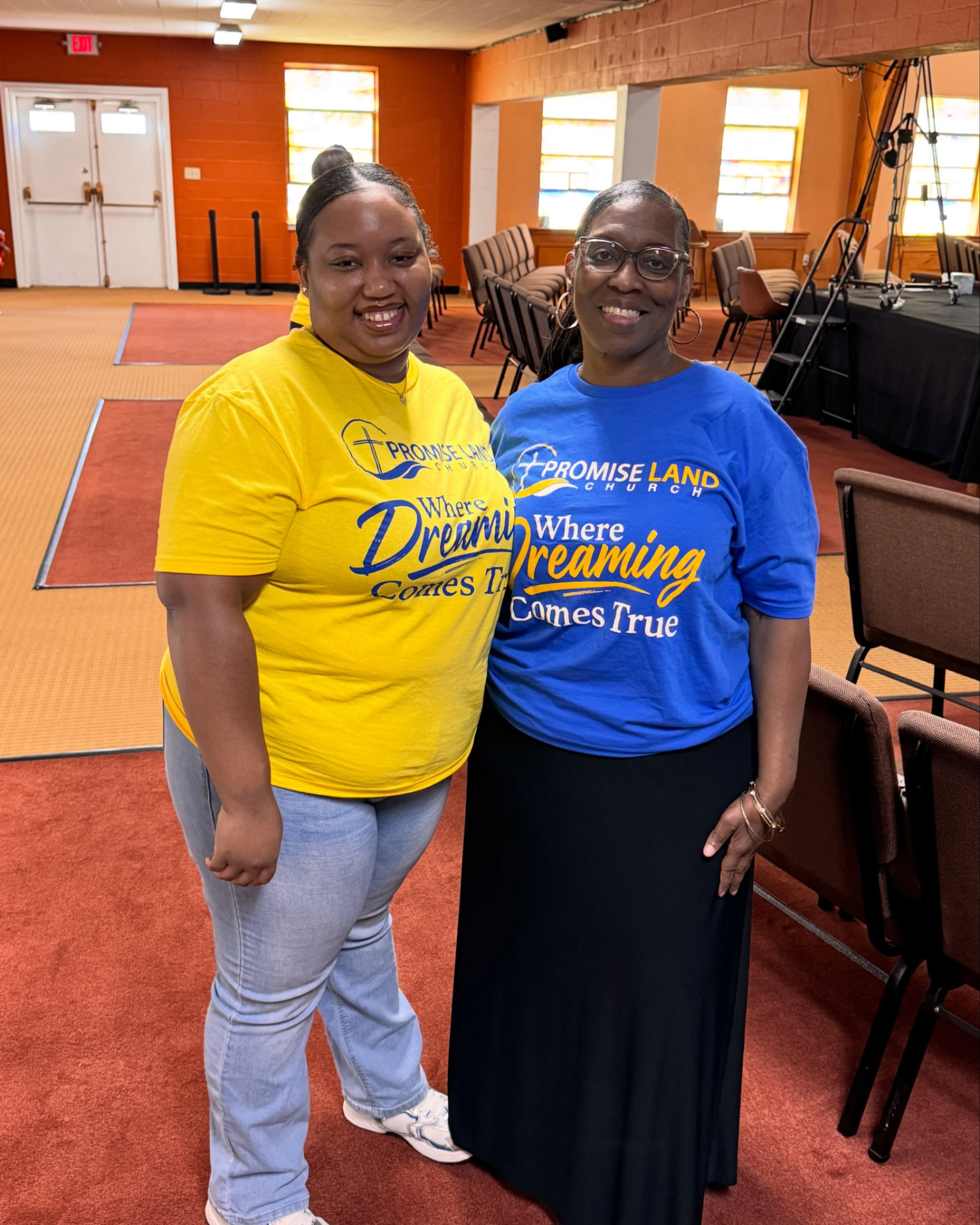 Two members in yellow and blue Promise Land Church 'Where Dreaming Comes True' shirts smiling together in the fellowship hall.